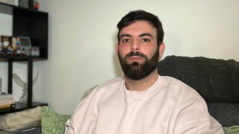 Igor sits on a sofa in a living room, wearing a light-coloured sweatshirt. Behind him is a black shelving unit with framed photos, books, small decorative objects, and a trailing plant against a pale wall.