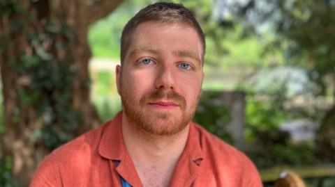 Headshot of Paddy Berryman, with a garden and tree in the background, he is wearing a coral coloured linen shirt