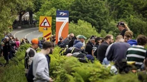 Fans line the side of the road at Hillberry on the the TT course. There is a road marker showing the section of the course as the road bends into the distance.