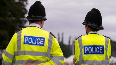 Two backs of two police officers can be seen. They are each wearing helmets and hi-vis jackets. It is a cloudy day.