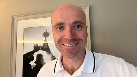 Andrew Lynch wears a white polo shirt with black lined collar and open neck. He is smiling at the camera and standing in front of a black and white image of a golfer holding up a cup.