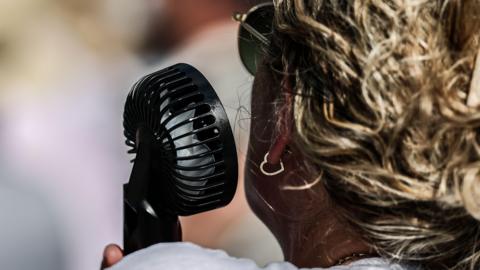 A close-up image of a black hand-held fan next to the left cheek of a woman, who is pictured from behind.