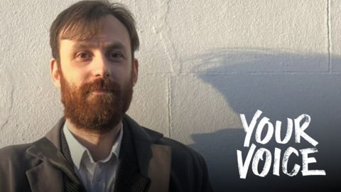 A man with brown hair and a beard smiling in the sun with a white wall background
