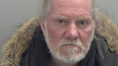 Graham Cockerill is seen in a police mugshot. He has receding white hair and a beard and he is glaring at the camera. He is wearing a dark puffer jacket with fur around the edge of the hood.