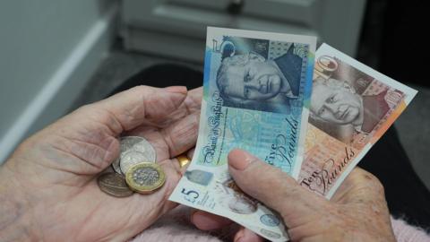 A five pound note, a ten pound note and coins are being held in the hands of an elderly person.