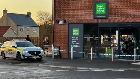 A lone police car parked outside a Co-op store. A police cordon tape is in place around the front of the store. The store is a red bricked building with a black entrance and some green signage. 