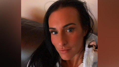 A selfie of Lisa Denton who has long dark hair and is slightly pouting into the camera. She is wearing a fluffy hoodie with dogs on them and sitting on a sofa.