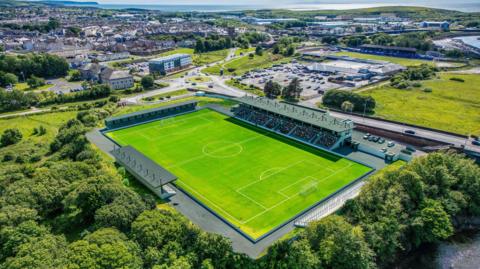A CGI image of what the new Cumberland Sports Village stadium would look like from the air. The grass pitch has a large stand along the far touchline which runs roughly penalty area to penalty area. At the far end and along the near touchline are smaller stands. A road runs behind the large stand, with the rest of the stadium surrounded by trees and green space.