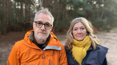 Doug Parr and Lynne Deeley standing in Sutton Heath woods. Doug is wearing an orange jacket and glasses. Lynne is wearing a navy jacket and a yellow coloured scarf.