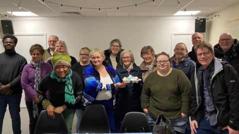 A group of 15 people stand in a room and smile at the camera. A woman in the middle of the group holds an Easter craft project.