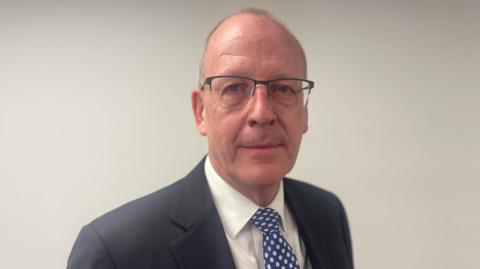 A man with very short grey hair and glasses looks towards the camera. He is wearing a grey suit jacket with a white shirt and blue tie with white spots