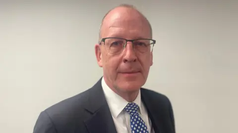 A man with very short grey hair and glasses looks towards the camera. He is wearing a grey suit jacket with a white shirt and blue tie with white spots