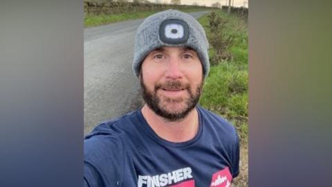 A man wearing a grey woollen hat and a blue T-shirt is smiling in a selfie. He has a brown beard. He is standing next to a road and a grass verge.