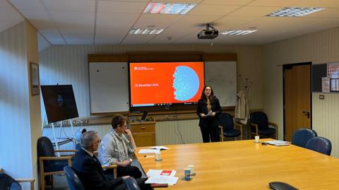 A room where Deputy Andrea Dudley-Owen is giving a presentation. The slide on show is red and has the words 25 November 2024 on it alongside a half completed semi-circular jigsaw. Two civil servants in smart attire are sat at a large wooden table. 