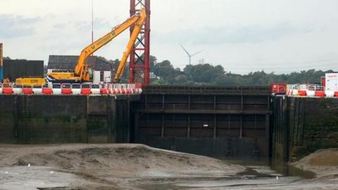 The closed sea gate at Glasson Dock, with a brown and green sea wall either side, the sandy earth of the dry dock in front and a yellow crane standing on the dock side