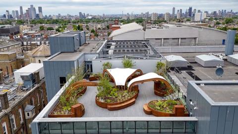 A hospital roof filled with flowerbeds and sweeping white canopies in the foreground, with other buildings and the London skyline visible in the background