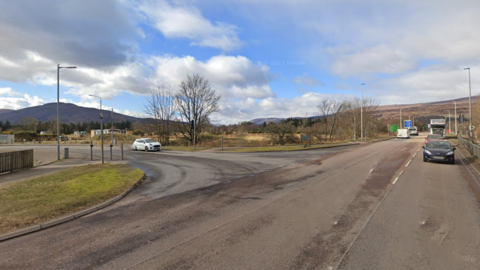 The area of the cycleway improvements in Fort William. There is a white car nearing a junction on the left and cars, lorries and vans on the road in front of it.