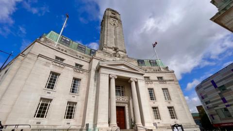A picture of the town hall in Luton, which is the headquarters of Luton Council. It is a grey building with four flours, and has a clock tower on top of it. The sky is a mixture of blue but also with some light grey clouds behind the building.