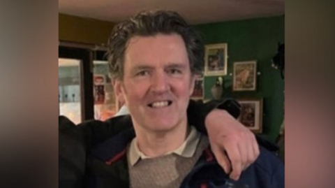 John McGarry is smiling. He has short brown hair and is wearing a grey jumper and a navy blue coat. Behind him is a wall with picture frames on it. The arm of someone out of shot is around his shoulders.