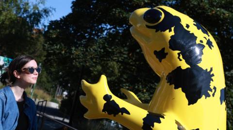 A large statue of a yellow and black frog-like creature with a woman in sunglasses looking at it