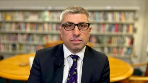A man with a black suit, white shirt and purple tie looking at the camera sitting in a library