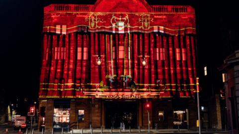 Video projection illuminates the façade of Edinburgh's King's Theatre at night, displaying colourful artwork across the historic building to mark its reopening following a major refurbishment. Traffic lights, pedestrians and the theatre entrance are visible in the foreground.