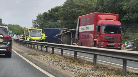 A lorry overturned on the A12. There are police officers standing around the car and there is an ambulance parked.