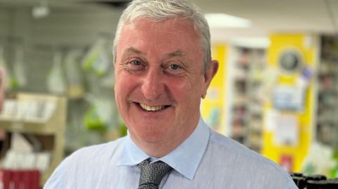 A man with light grey hair smiles at the camera. He is wearing a light blue shift and a dark grey tie. He stands inside a chemist.