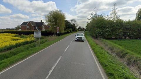 A road, flanked by fields, with a black on white sign stating the name of the village - Shepeau Stow. 