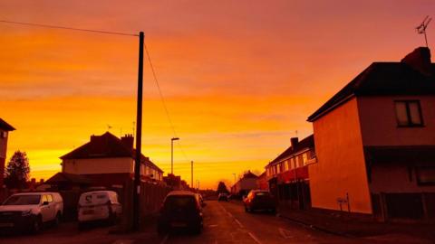 An orange and yellow sky over houses. Several cars are parked on the residential streets and covered in the warm glow.