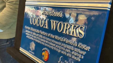 A rectangle blue plaque reads:
Rowntree's Cocoa Works. Former Chocolate Factory of the world-famous KitKat. between the 1890s and 2006, this factory pioneered progressive workers' welfare for the tens of thousands who passed along this path, sharing a strong sense of friendship and community through work and leisure.