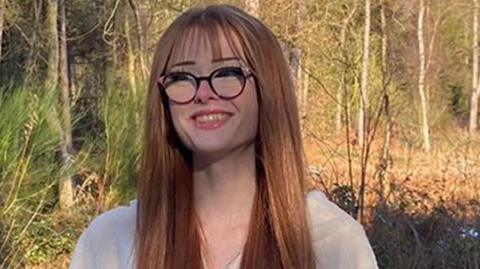 Brianna Ghey, who has long straight brown hair and wears black glasses, smiles at the camera while standing against a backdrop of woodland