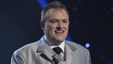 A white man with brown hair, who seems to be in his forties. He is smiling, looking past the camera. The background is dark with some strobe type lighting - he is on stage. He is wearing a satin-looking light grey suit, white shirt, and black tie. He is holding a microphone.