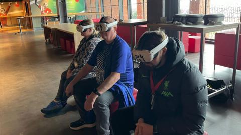 Two men and a woman, wearing white virtual reality headsets, are shown sitting on padded stools.