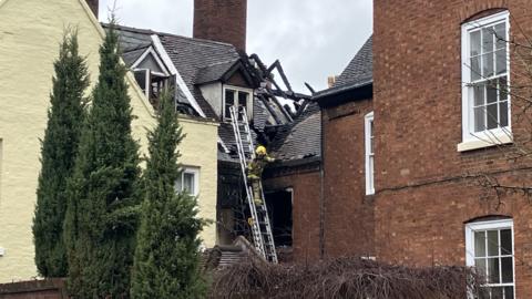 A firefighter with a yellow helmet on a ladder against a property. The roof beams are exposed after a fire and they are charred.