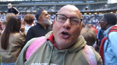 A man wearing a khaki green hoodie, wearing a pink rucksack and black-framed glasses, appears to talk or shout towards the camera. Crowds are behind him on the MK Dons football pitch, with the stands in the background.