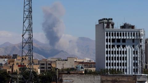 Smoke rises in the mountains in the distance as we look at building tops from far away in Iran as well as a metal tower.