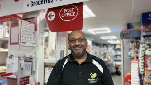 A man in a black polo shirt in a shop, underneath a red sign with the Post Office logo. The walls around him contain shelves full of goods or are covered in information leaflets.