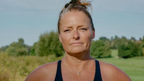 A head and shoulders photo of Laura Reineke, a woman with blonde hair tied in a bun. She's wearing a black swimming costume and a necklace with a mermaid on. She's looking straight at the camera with a half-smile, with trees and a blue sky behind her.