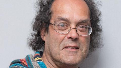 Head and shoulders shot of Michael Pascal who is balding with dark curly hair and wearing metal-rimmed glasses