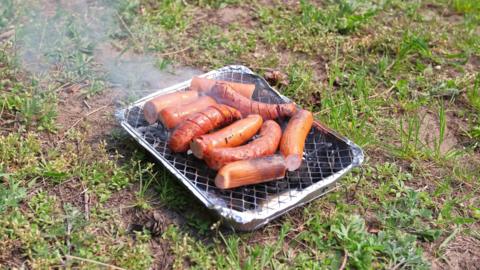 Sausages sizzling on a disposable barbecue. The barbecue is on a patch of grass.