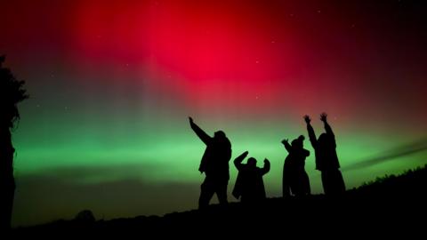 Silhouetted figures raise their arms toward the sky beneath a striking display of red and green aurora lighting the night.