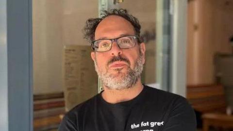 Andy Sparsis stands in front of his restaurant. He wears glasses and a black t-shirt.