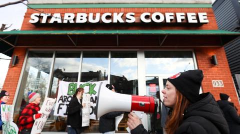 A young female union leader in a black and red woollen hat is standing among other picketers outside a Starbucks. Some of the other picketers in the background are holding signs whose message is not visible in the image. The female union member in the foreground is holding a megaphone as if she is about to start speaking into it.