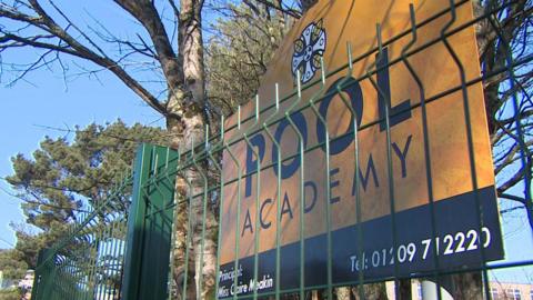 The image shows a sign for Pool Academy behind a green metal fence, with trees and school buildings visible in the background. It appears to be a daytime exterior shot of the school's entrance on a clear, sunny day.