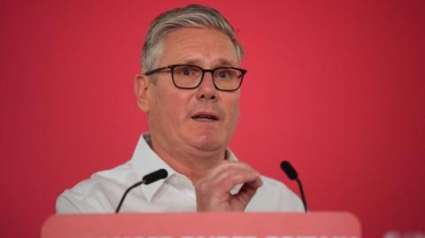 Starmer gestures with right hand while speaking in front of two microphones
