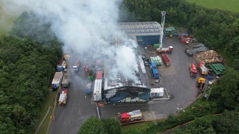 A drone shot of smoke coming out of the roof a large corrugated iron industrial building, surrounded by several fire fighting vehicles in a car park. The car par is surrounded by trees.