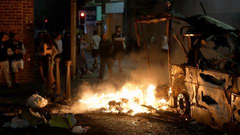The carcass of a vehicle is on fire at night. Crowds gather around.