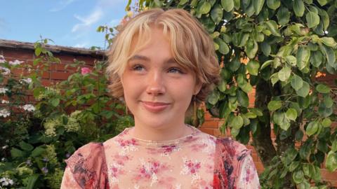 Close up of Niamh Shergold. She is stood outside in front of a red brick wall with green trees and shrubbery. She has short light hair and is wearing a pink floral top.