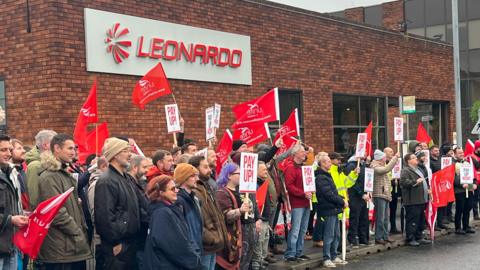 A large group of Leonardo workers waving red flags promoting Unite the union. They are standing outside the Yeovil factory and most are wearing coats and hats. 